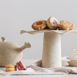 On the right is a cake stand with cookies on top. On the left is a teapot of the same material. They are made of mottled beige stoneware. On the table, there are several sweets.  Light mood photography.