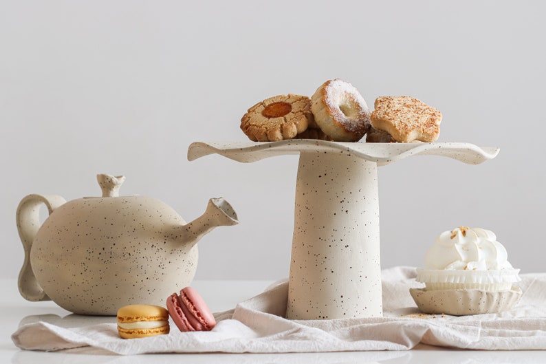 On the right is a cake stand with cookies on top. On the left is a teapot of the same material. They are made of mottled beige stoneware. On the table, there are several sweets.  Light mood photography.