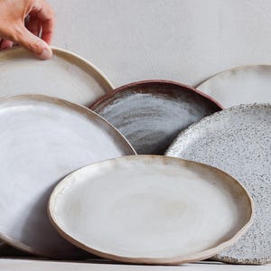 Könnte beinhalten: Eine Sammlung handgefertigter Keramikplatten in verschiedenen Größen und Farben. Die Teller weisen eine Reihe von neutralen Tönen auf, darunter Weiß, Beige und Grau, wobei einige gesprenkelte oder strukturierte Oberflächen aufweisen. Die Teller sind in einer geschichteten Anordnung angeordnet.