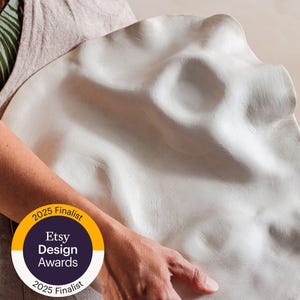 Pode incluir: Um prato de cerâmica grande, branco, com formato orgânico. O prato é segurado por uma pessoa que usa um avental de linho. Um adesivo no avental indica que o item é um finalista do Etsy Design Awards 2025.