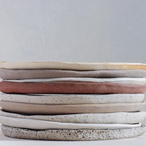 Op de afbeelding: Een stapel van negen keramische borden met onregelmatige, handgevormde randen. De borden hebben diverse aardetinten, waaronder crème, taupe, terracotta en gespikkeld wit, met uiteenlopende texturen en matte afwerkingen in een verticale opstelling.
