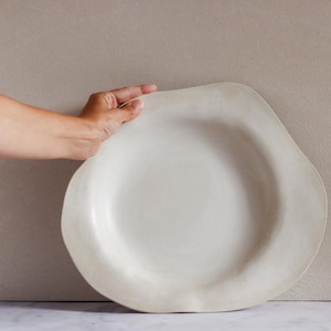 Puede incluir: Un plato grande de cerámica blanquecina con una forma orgánica e irregular. El plato tiene un borde ancho y una superficie ligeramente texturizada. La mano de una persona sostiene el plato, lo que sugiere su tamaño y forma. El plato es probablemente para servir comida.