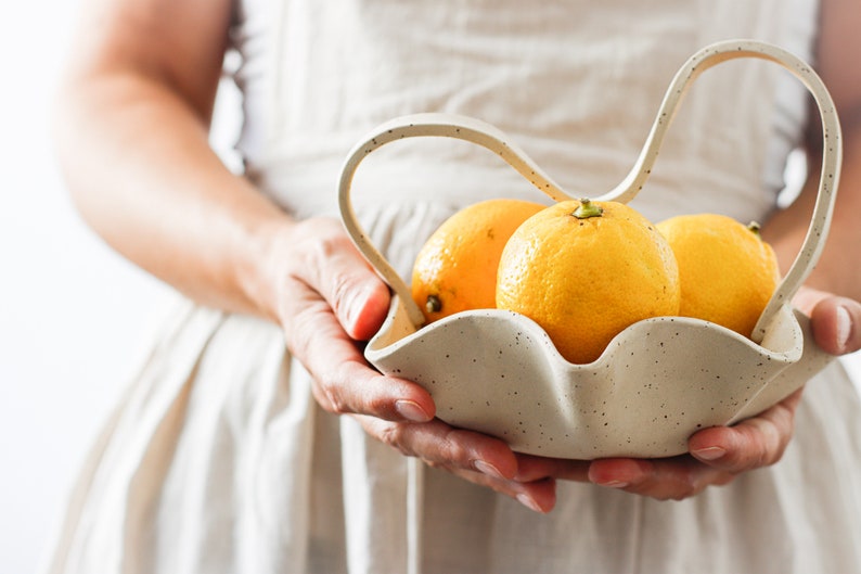 The torso of a woman in an apron is seen holding a wavy basket of mottled beige stoneware with a wavy handle. It contains lemons.