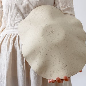 A woman in a beige apron holds a large ceramic fruit bowl. It is mottled beige and wavy in shape. It shows the back, where an author's stamp can be seen.
