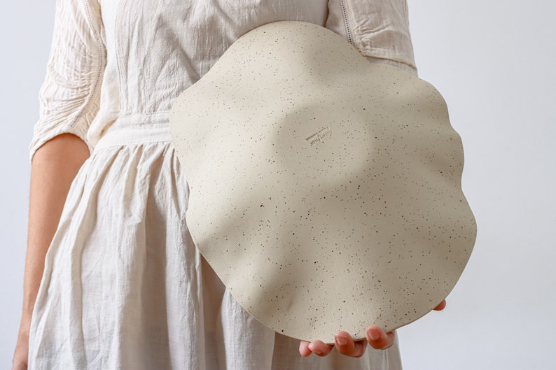 A woman in a beige apron holds a large ceramic fruit bowl. It is mottled beige and wavy in shape. It shows the back, where an author's stamp can be seen.