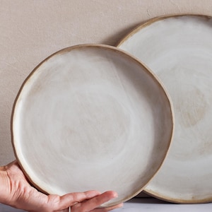 Puede incluir: Dos platos de cerámica blanquecina con superficie texturizada y borde ligeramente elevado. Los platos son redondos, uno de ellos sostenido por una mano. Los platos son de color neutro, adecuados para diversos entornos de comedor.