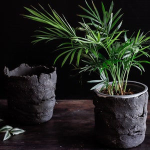 On a dark table, two utensil holders are facing each other. One contains a plant. The utensil holders are made of matte black clay with a lot of texture. They are made with strips of clay joined randomly.