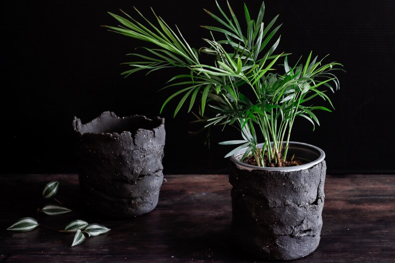 On a dark table, two utensil holders are facing each other. One contains a plant. The utensil holders are made of matte black clay with a lot of texture. They are made with strips of clay joined randomly.