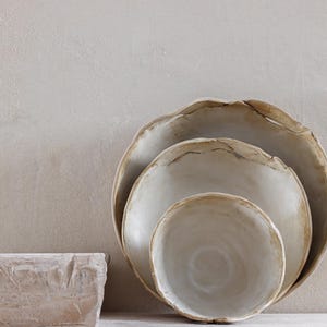Puede incluir: Una pila de tres cuencos de cerámica blanquecina con bordes irregulares y ondulados. Los cuencos están anidados, el más grande en la parte posterior y el más pequeño en la parte delantera. Un objeto rectangular y texturizado está en primer plano.