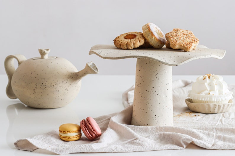 Still life in high-key. On the right is a beige marbled stoneware cake stand with several cookies on top. On the table, there is a beige cloth with several sweets on top. On the left is a teapot of the same material.