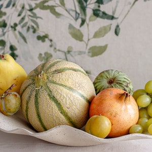 May include: A white ceramic bowl with a wavy rim holds a variety of fruits, including a yellow quince, a green melon, a green apple, a green and white grape cluster, and a bright orange pomegranate.