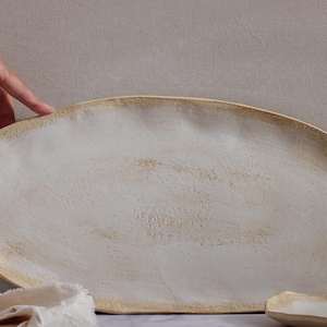 One hand rests lightly on the edge of a very large oval slab of matte white glazed ceramic.