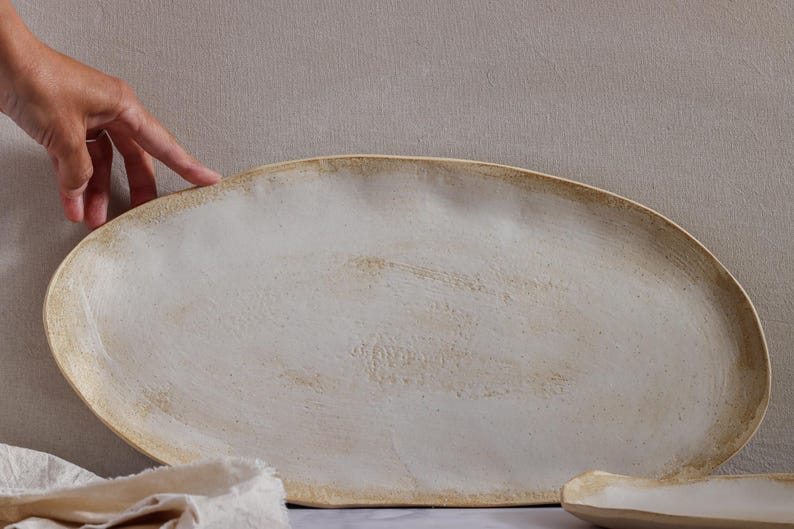 One hand rests lightly on the edge of a very large oval slab of matte white glazed ceramic.