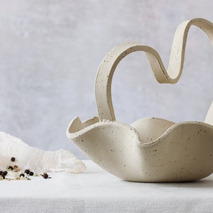 A mottled beige ceramic basket on a table with a white tablecloth. It has wave-shaped edges, and the handle is in an M-shaped curve. On the left can be seen a glass salt grinder.