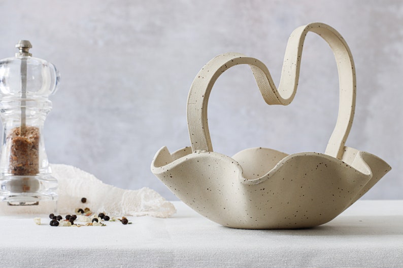 A mottled beige ceramic basket on a table with a white tablecloth. It has wave-shaped edges, and the handle is in an M-shaped curve. On the left can be seen a glass salt grinder.