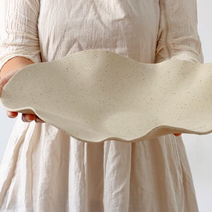 A woman holds a very long fruit bowl in the shape of waves. It is stoneware and mottled beige in color.