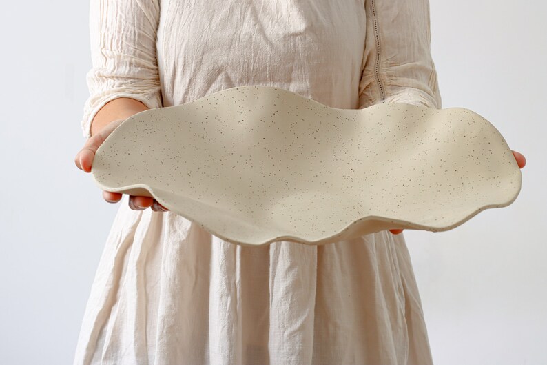 A woman holds a very long fruit bowl in the shape of waves. It is stoneware and mottled beige in color.