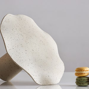 A cake stand rests horizontally on a white table so that the top of the cake stand can be seen. It is made of beige marbled stoneware and has waves on the edges of the tray. Next to the cake stand are several macarons.