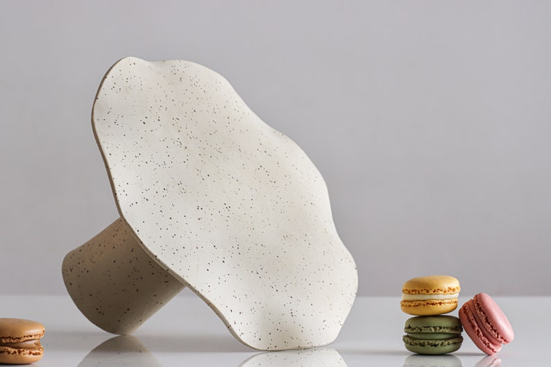 A cake stand rests horizontally on a white table so that the top of the cake stand can be seen. It is made of beige marbled stoneware and has waves on the edges of the tray. Next to the cake stand are several macarons.