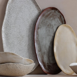 Against a wall is an oval terracotta tray, a larger black and white one to its left, and a smaller beige one to its right. To its left, on a table, are two handmade stoneware soup bowls.