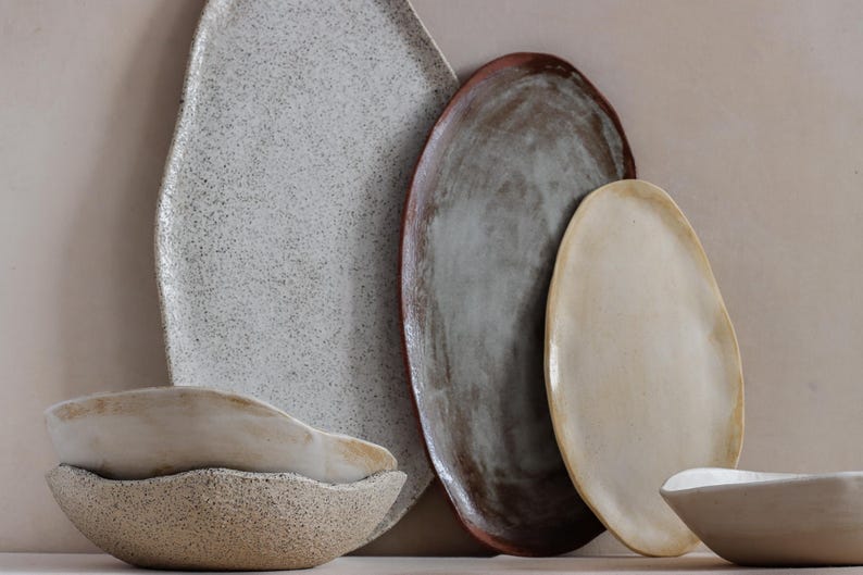 Against a wall is an oval terracotta tray, a larger black and white one to its left, and a smaller beige one to its right. To its left, on a table, are two handmade stoneware soup bowls.