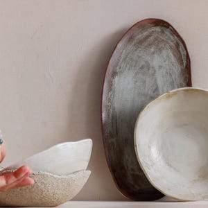 Against a wall, an oval terracotta tray and a deep dish leaning forward, revealing its contents. Next to it, on a table, a hand lifts a deep stoneware ceramic dish.