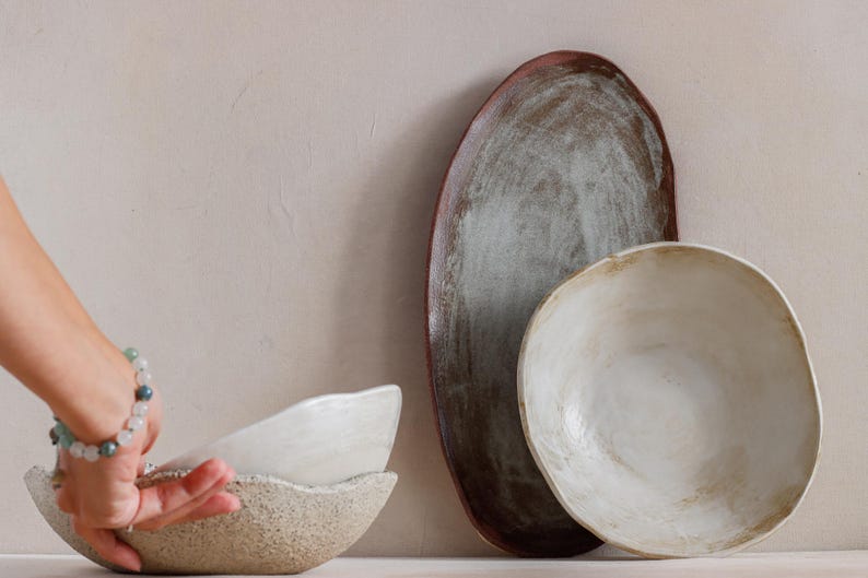 Against a wall, an oval terracotta tray and a deep dish leaning forward, revealing its contents. Next to it, on a table, a hand lifts a deep stoneware ceramic dish.