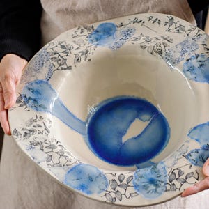 Może przedstawiać: Duża biała ceramiczna miska z niebieską glazurą. Miska ma szeroki rant z ozdobnym wzorem czarnych i niebieskich wzorów kwiatowych.