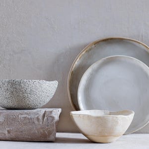 Può includere: Una collezione di stoviglie in ceramica fatte a mano. L'immagine presenta una ciotola grigia strutturata, una piccola ciotola bianco sporco e due piatti nei toni del grigio e del beige. I piatti hanno un'estetica rustica e organica.
