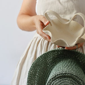 A woman holds a mottled beige ceramic basket with a wavy handle in the shape of an M. She also holds a large country green hat.
