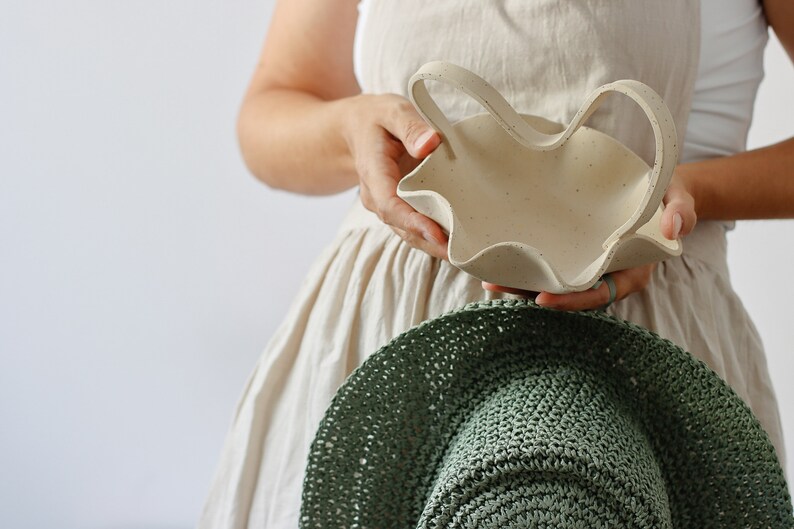 A woman holds a mottled beige ceramic basket with a wavy handle in the shape of an M. She also holds a large country green hat.