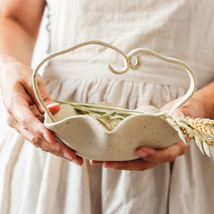 A woman holds a dotted beige ceramic basket in the shape of waves. It has a handle formed by two stoneware strips of the same color as the basket, which are joined in the center forming two small spirals. The basket contains spikes.