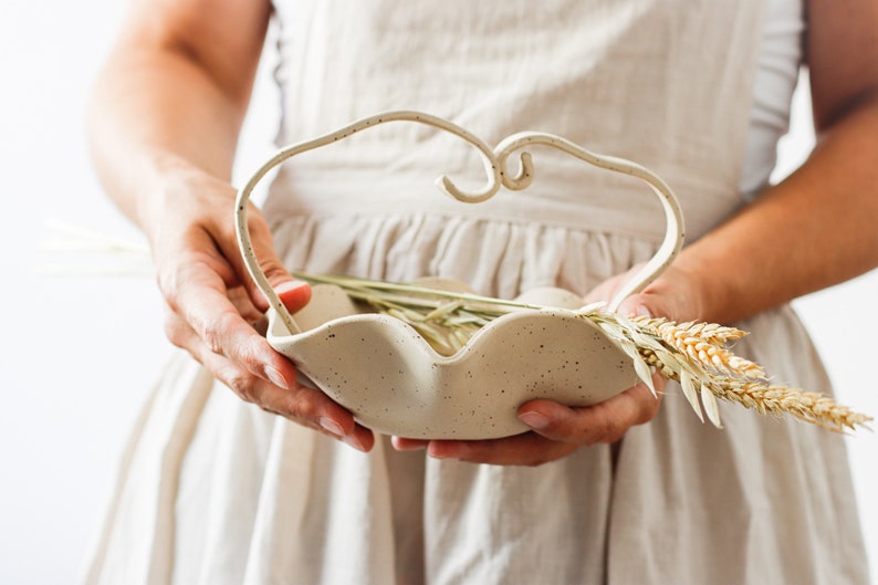A woman holds a dotted beige ceramic basket in the shape of waves. It has a handle formed by two stoneware strips of the same color as the basket, which are joined in the center forming two small spirals. The basket contains spikes.