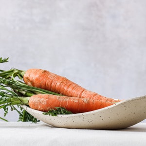 On a white tablecloth is an oval bowl that can be seen almost in profile. It is of mottled beige clay and contains two carrots with their foliage. Gray background.
