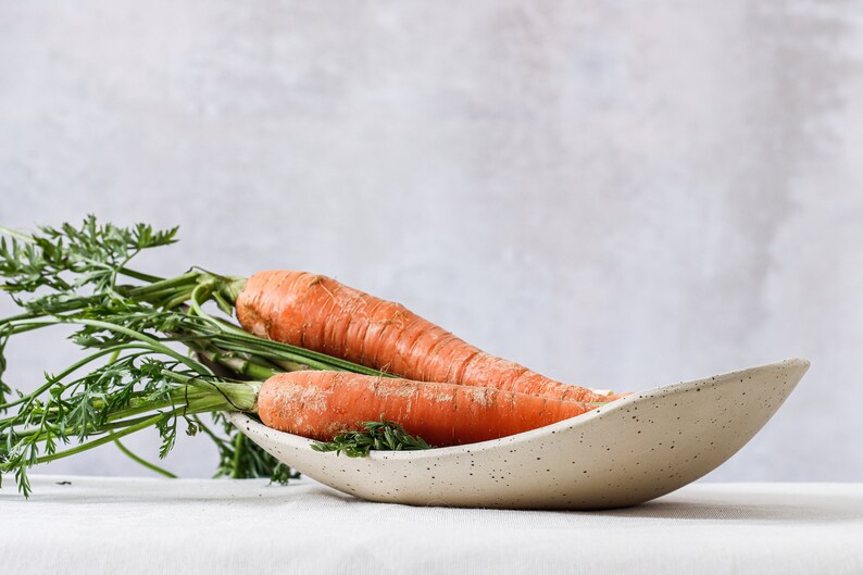 On a white tablecloth is an oval bowl that can be seen almost in profile. It is of mottled beige clay and contains two carrots with their foliage. Gray background.