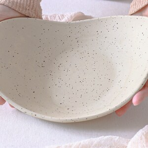 View at 45º. Two hands hold an oval fruit bowl in mottled beige clay. The fruit bowl is deep, and a light pink cloth completes the scene.