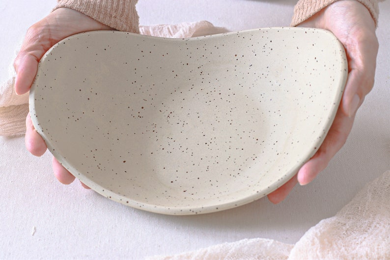 View at 45º. Two hands hold an oval fruit bowl in mottled beige clay. The fruit bowl is deep, and a light pink cloth completes the scene.