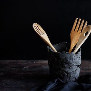 A black vessel made from strips of hand-cut, textured clay. It has holes made with the fingers. It is on a wooden table and the bottom is black. It contains three wooden spoons. A piece of cloth is seen on which the utensil holder.