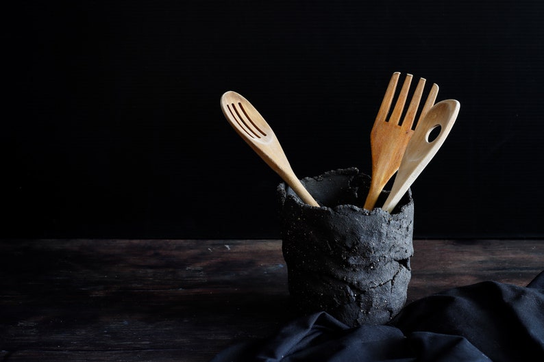 A black vessel made from strips of hand-cut, textured clay. It has holes made with the fingers. It is on a wooden table and the bottom is black. It contains three wooden spoons. A piece of cloth is seen on which the utensil holder.