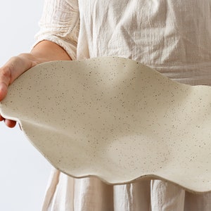 A woman in a beige apron holds a very long centerpiece in the shape of waves. It is beige and made of ceramic.