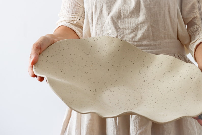 A woman in a beige apron holds a very long centerpiece in the shape of waves. It is beige and made of ceramic.