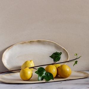 On a marble table is an oval ceramic bowl with lemons on top. Behind it is a similar fountain leaning against the wall. It is made of matte white stoneware