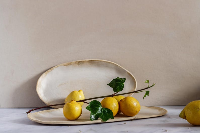On a marble table is an oval ceramic bowl with lemons on top. Behind it is a similar fountain leaning against the wall. It is made of matte white stoneware