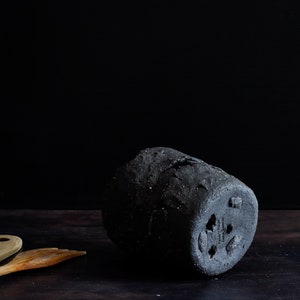 Dark Mood photography of still life. The back of a utensil holder can be seen. It has three holes and three small legs. Holes are made by hand. Next to it on the wooden table are two wooden spatulas. Black background.