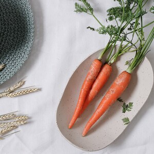 Zenithal view of an oval fruit bowl in mottled beige clay. It contains several carrots with their foliage. On the left can be seen a part of a green hat and some ears of corn.