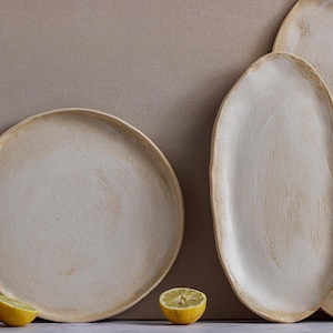 A plate is seen from the front leaning against a wall, and next to it are two oval dishes in matte white glazed stoneware.