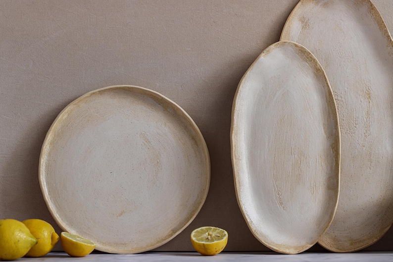 A plate is seen from the front leaning against a wall, and next to it are two oval dishes in matte white glazed stoneware.