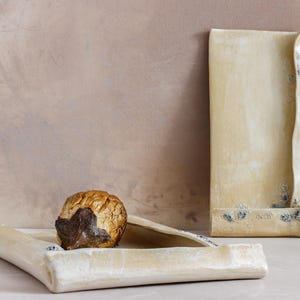 May include: A still life composition featuring handmade ceramic pieces. The image showcases a textured, beige-colored rectangular tray, a tall, folded ceramic piece, and a decorative object with a brown and gold exterior.