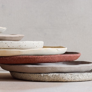 Può includere: Una pila di piatti in ceramica fatti a mano in vari colori e trame. I piatti includono sfumature di bianco, beige, crema macchiato, terracotta e grigio. I piatti sono impilati, mostrando le loro forme e finiture uniche.
