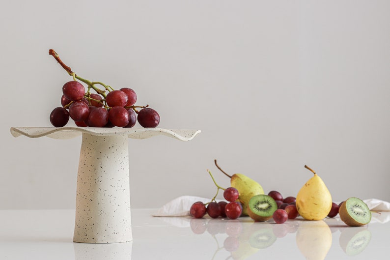 Still life scene in high-key. On the left is a tall cake stand, with a cone-shaped base. The upper part has waves. On top is a bunch of red grapes. Behind it are various fruits such as kiwis, grapes, and pears.